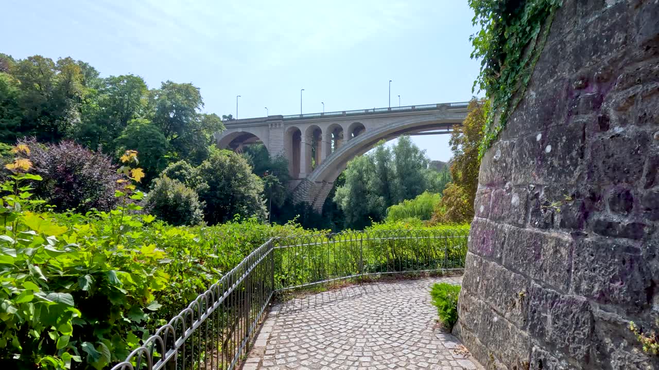 Camera moves along a cobblestone path bordered by greenery, revealing a large stone arch bridge in a sunlit European park setting