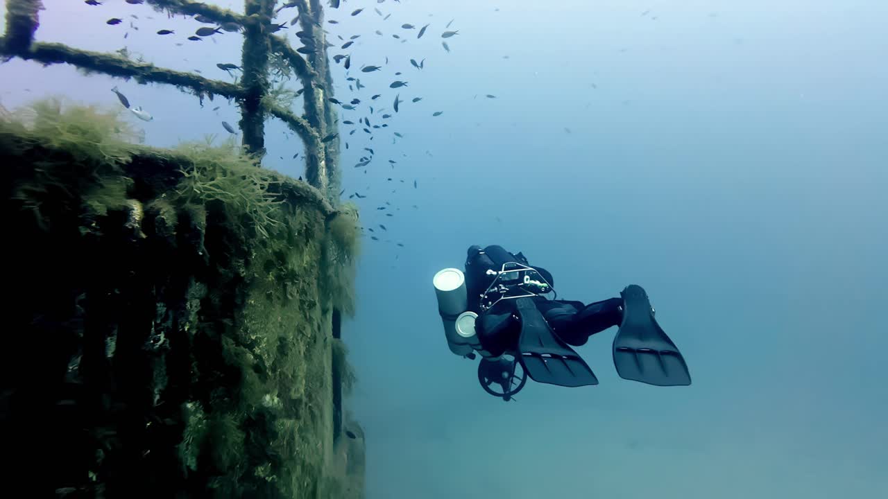 Subacqueo tecnico con scooter subacqueo esplora il ponte metallico e le ringhiere muschiose di un relitto affondato nel profondo Mar Mediterraneo blu al largo di Malta e Gozo durante un'immersione di un soleggiato giorno d'estate.
