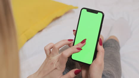 woman holding a smartphone with green screen on a bed