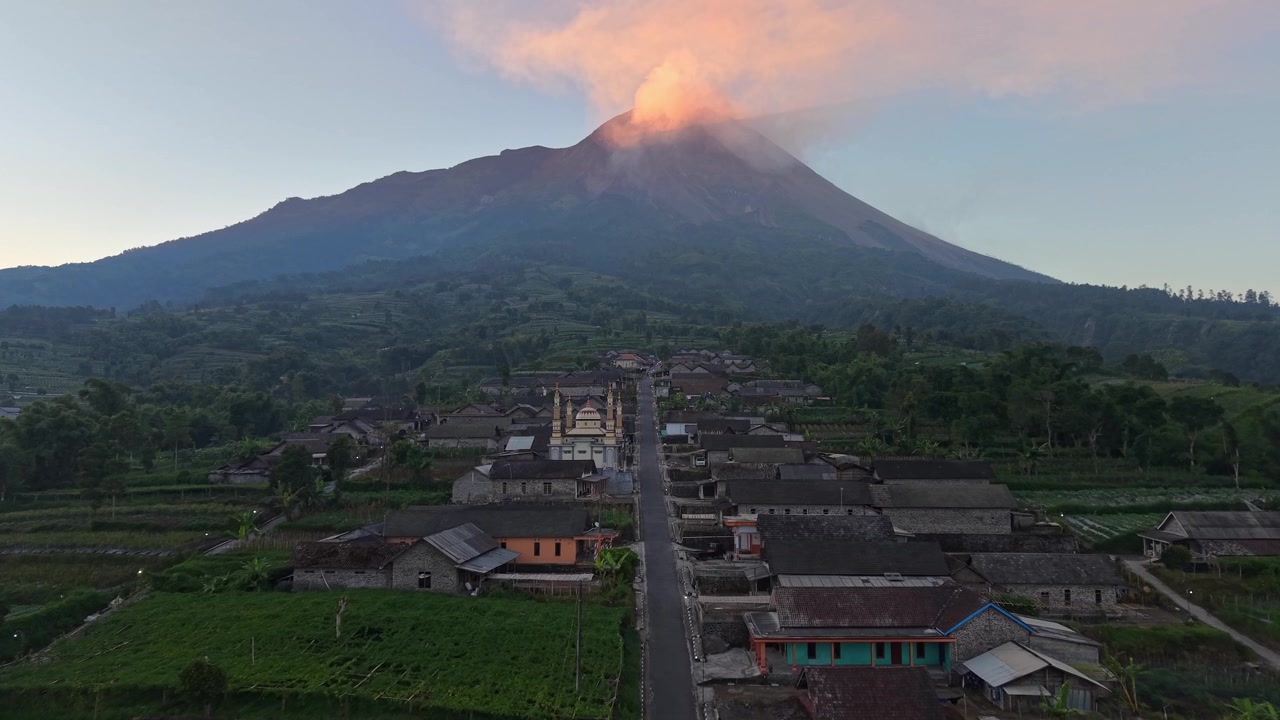 Veduta aerea di un paesaggio con il Monte Merapi che si erge sopra una comunità rurale. Vegetazione verde, terreni agricoli e drammatiche nuvole vulcaniche all'alba.