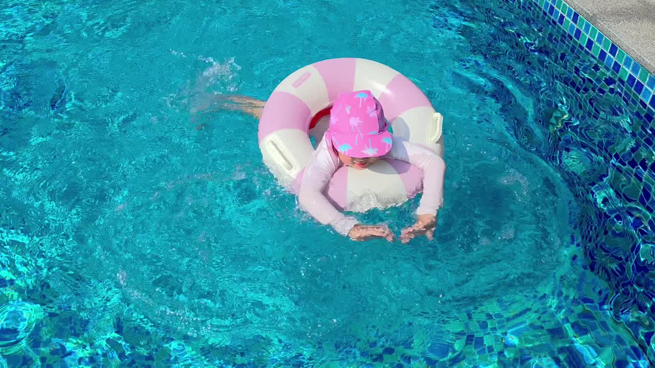 Tracking shot following a 5-year-old girl wearing a pink hat and floatie ring as she learns to swim by kicking her legs in a deep, blue swimming pool on a sunny day