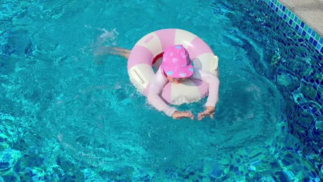 tracking shot following a 5-year-old girl wearing a pink hat and floatie ring as she learns to swim by kicking her legs in a deep, blue swimming pool on a sunny day