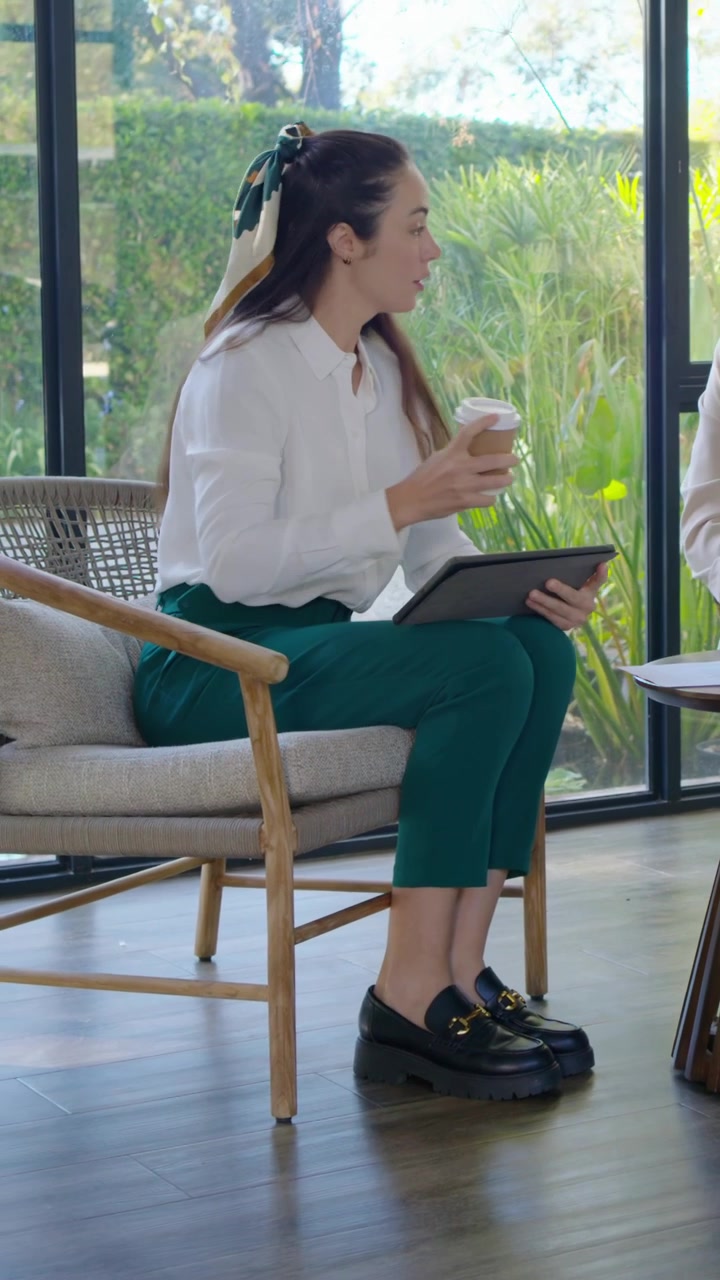Vertikal video: Kvinde i bluse læner sig, retter sig til i lounge, tager en kop, taler og lægger mærke til en tablet.
