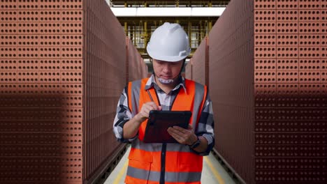 asian male engineer with safety helmet taking note on the tablet and looking around while standing with red brick packed in stacks are stored