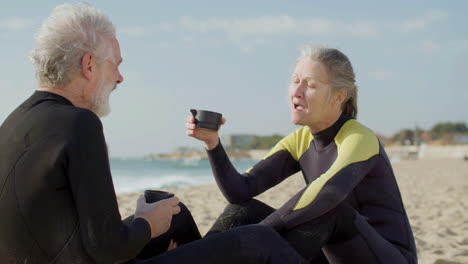 senior couple in wetsuit drinking tea and talking together while sitting on a surfboard at the beach 1