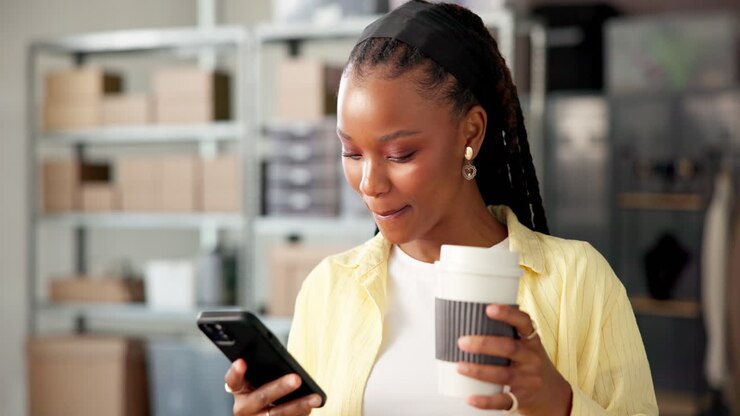 Young Black Woman Using Phone and Drinking Coffee in Warehouse