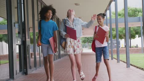 Video-of-three-diverse-schoolgirls-walking-in-school-corridor-talking-and-laughing,-copy-space