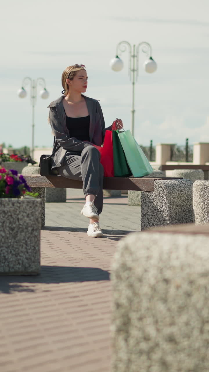 dama blanca sentada en un banco al aire libre con bolsas de compras de colores a su lado, mirando hacia la distancia, las flores cercanas revolotean en el viento mientras se pueden ver postes de luz en el fondo