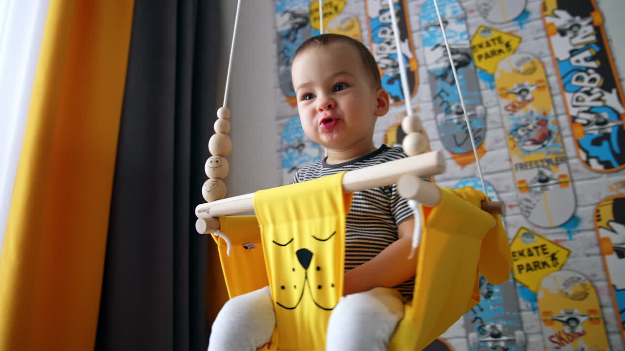 Active cute Caucasian baby boy sitting in a yellow swing. Happy kid swinging at home.