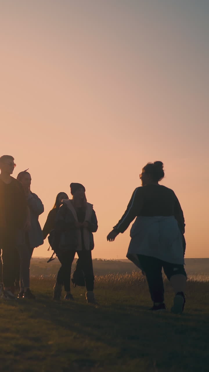 group of joyful friends walks along river bank to modern camp with tent in calm autumn evening at back sunset on horizon