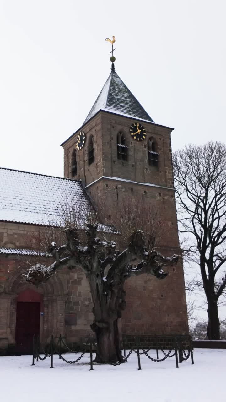 Historisk kirketårn dekket av et lett snølag, som står mot en blek vinterhimmel. Nakne trær rammer inn den gamle steinbygningen, og viser frem dens arkitektoniske detaljer og tidløse skjønnhet i