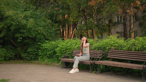 woman sitting on wooden bench outdoors, holding phone in her right hand while adjusting her hair with her left, surrounded by lush greenery and trees, with building