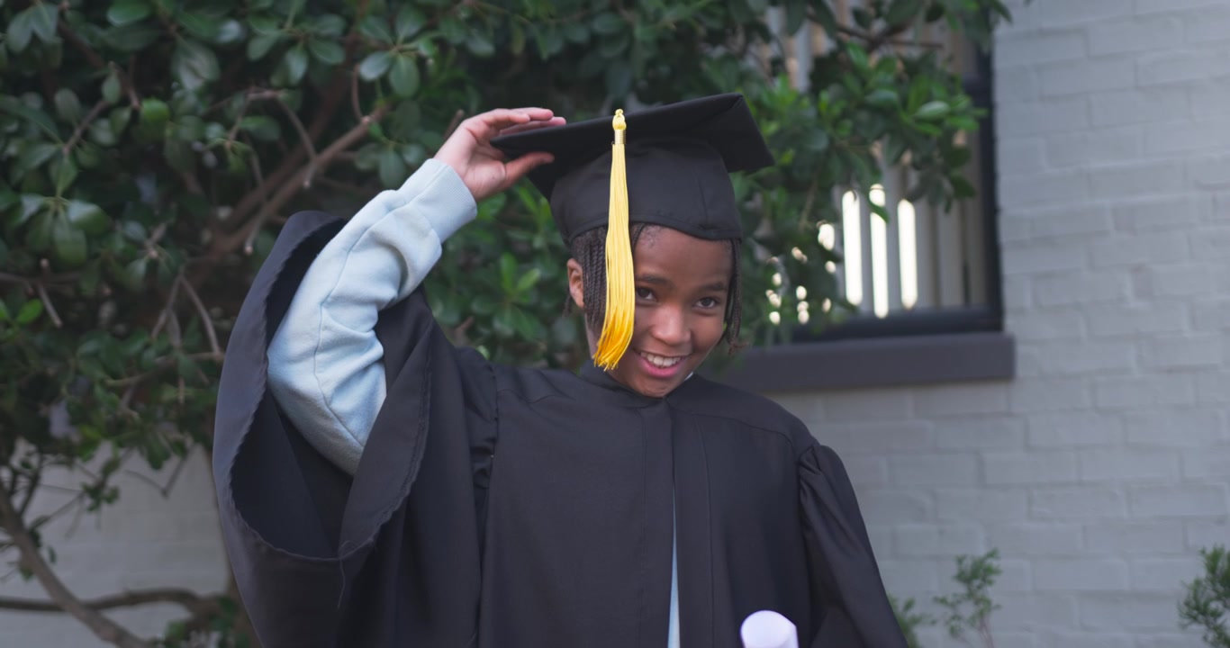 African American youth in black gown lifting cap then tossing with diploma for graduation at home
