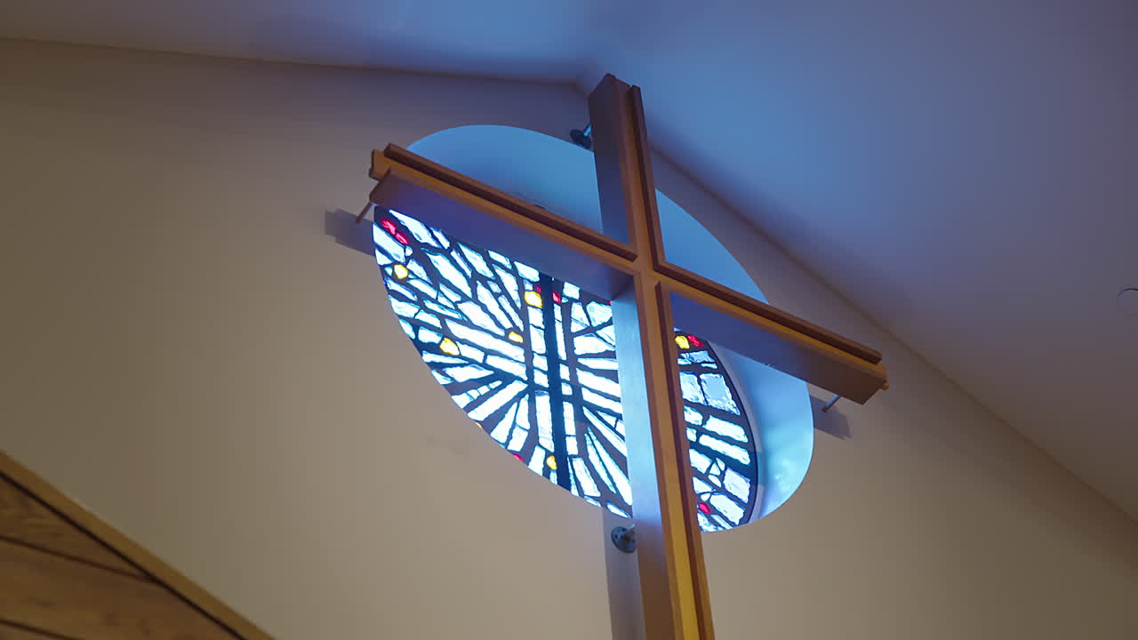 A tall wooden cross stands before a circular stained glass window in a quiet church sanctuary