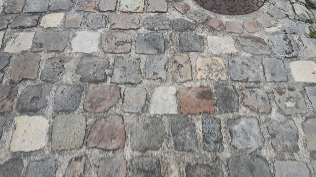 Low angle tracking shot over historic granite cobblestones and a vintage metal manhole cover