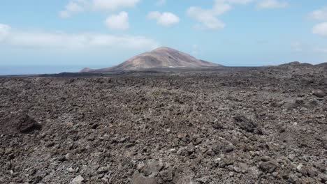 Drohne-Fliegt-über-Trockenes-Lavafeld-Mit-Einem-Vulkankrater-Im-Hintergrund,-Insel-Lanarote,-Sonniges-Wetter