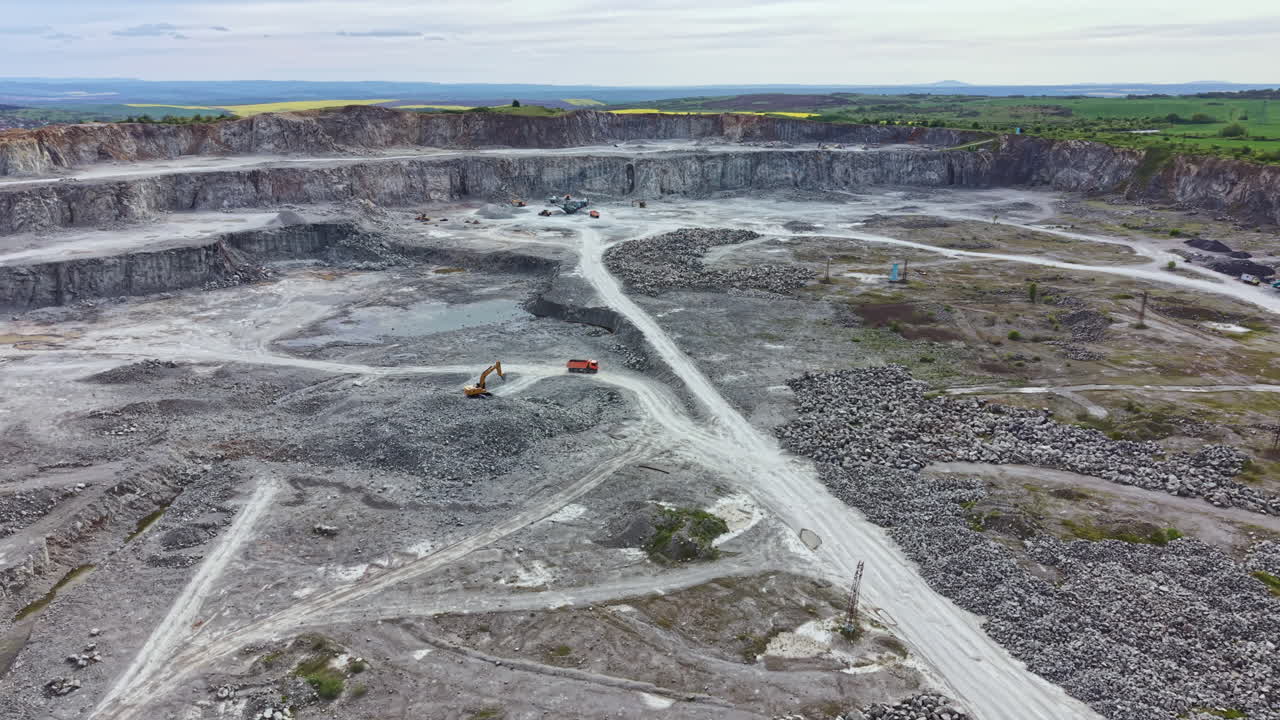 Mining operation takes place in a large open pit with machinery working on a cloudy day in a remote area with hills in the background
