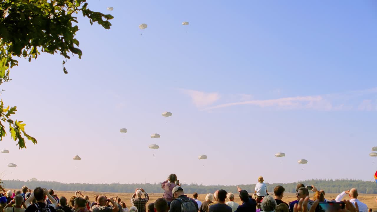 Large crowd watches as military parachutists descend in formation against a clear blue sky. The audience, including families with children, captures the moment with cameras during a mass drop