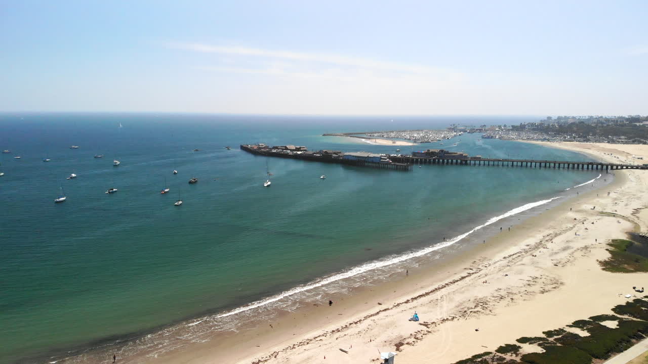 tiro aéreo cayendo sobre la playa de arena con vistas al océano azul y al muelle de madera de stearn's wharf en santa barbara, california