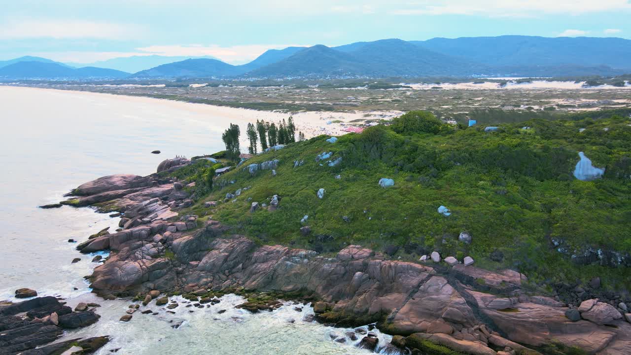 Joaquina Beach seen from above, an aerial view of the rocky coastline with Atlantic Forest and turquoise ocean with many mountains and dunes on the horizon in Florianopolis, Santa Catarina, Brazil