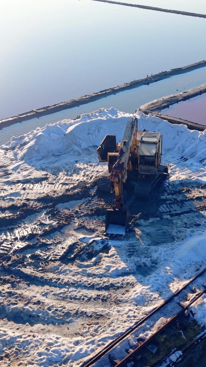 Excavator scooping salt mound, Bulgaria. Aerial view of machine amid white pile with blue channel and pink water beyond. Vertical video.