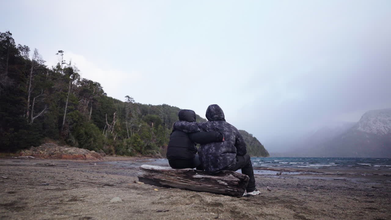 Two subjects huddle together on lake shore log, with their hug showing silent defiance of the cold outdoors. Ideal for friendship-love stories or survival bond narratives