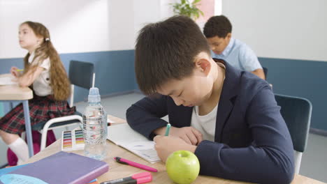 학교 영어 수업 시간에 책상에 앉아 공책에 글을 쓰는 소년