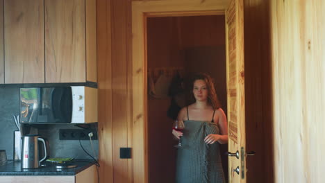 women wrapped in towels holding wine glasses walking through wooden doorway from rustic kitchen, illuminated by warm soft light, expressing relaxed casual mood as they move in cabin environment