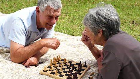 Älteres-Paar-Entspannt-Im-Park,-Liegt-Auf-Einer-Decke-Und-Spielt-Schach