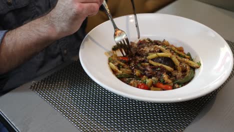 man eating a pasta dish with a fork and knife on a grey table