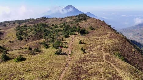 Aerial view of hikers trekking along scenic mountain ridge trail with a majestic mountain in the background. Clear blue sky and natural landscape highlight of outdoor adventure. Mount Prau, Indonesia