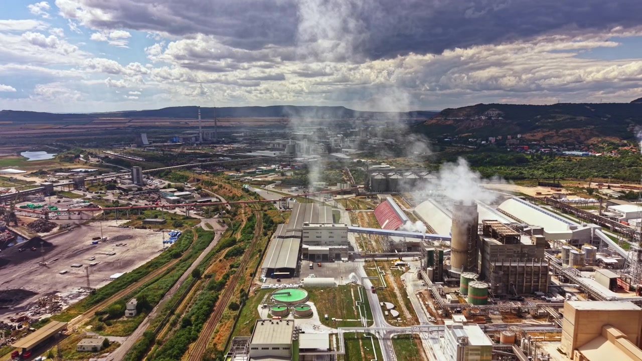 Industriområde med fabrikker og røyk som stiger opp mot himmelen nær en by på dagtid i et solrikt miljø