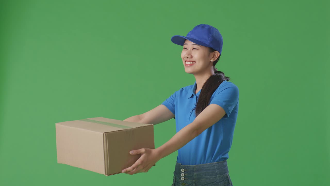 Asian Female Courier In Blue Uniform Giving A Carton To Someone And Smiling While Delivering It On Green Screen Background In The Studio