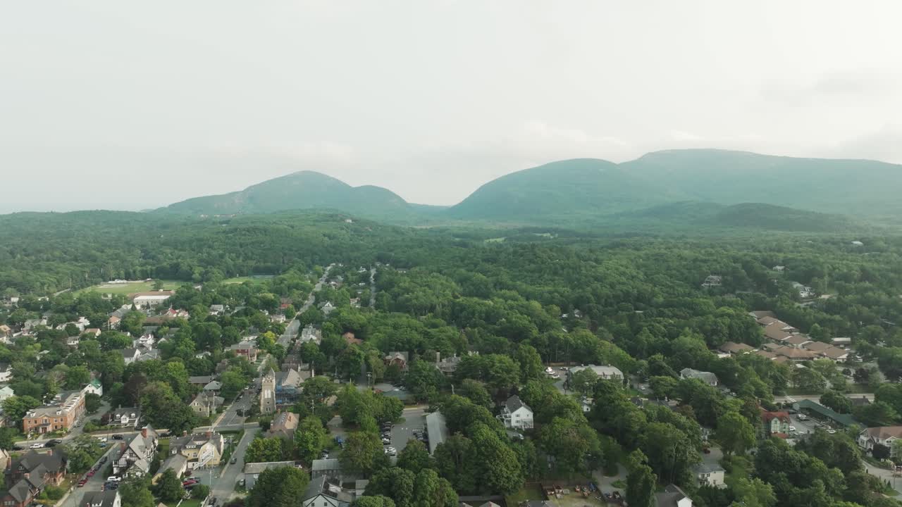 Drone view of Bar Harbor, Maine with coastal town charm and Acadia’s forested mountains in the backdrop on a hazy summer day.