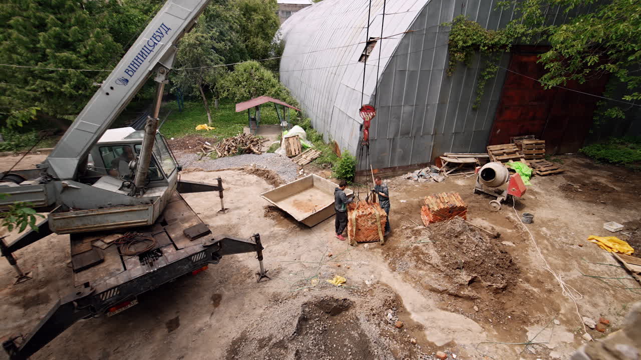 Two men attach the pile of red brick pile to the hook of an excavator. A look at the yard of the building under construction.