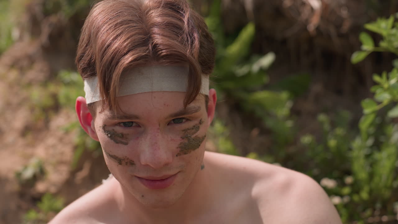 brown hair warrior wearing headband and mud face paint sharply turns toward camera, extended thumb and index finger shaped like gun, intense gaze and battle mood on rugged riverbank under daylight