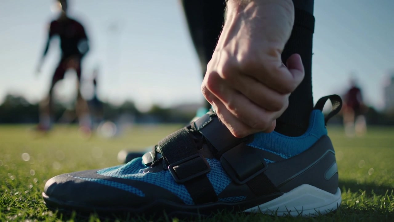 Mano che si allunga per regolare i lacci blu della scarpa da calcio dell'atleta e il calzino nero sul campo in erba sintetica, preparandosi per l'allenamento