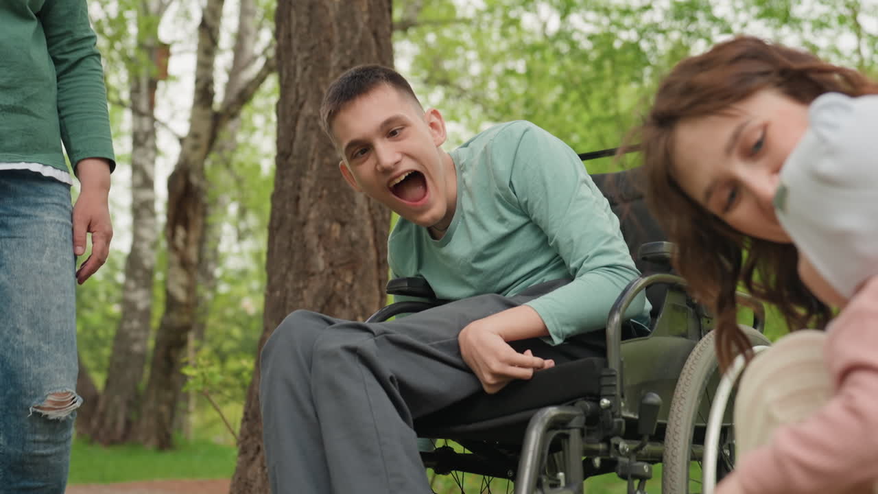 Hombre blanco en un parque en silla de ruedas riendo mientras una madre ayuda a su bebé a dar sus primeros pasos con delicadeza. Una cuidadora y la madre comparten una reacción alegre. Árboles primaverales, ambiente integrador y esperanzador