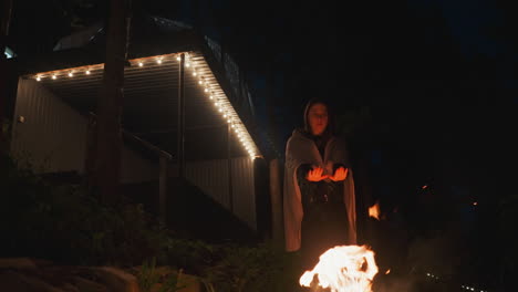 dark woman silhouette warming hands over bonfire at night. lady with blanket on shoulders trying to keep warm in cold evening. walk in courtyard of glamping house