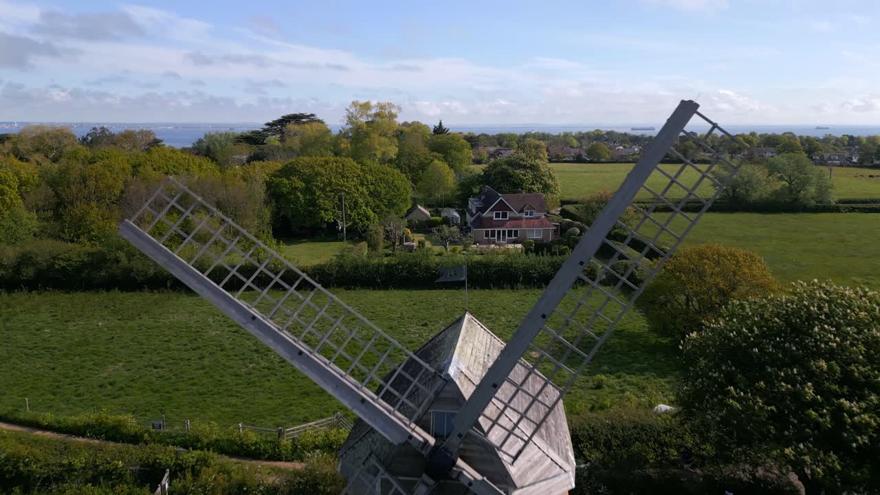 Forward drone shot flying between Bembridge Windmill blades, rising to reveal the stunning coastline, sea views, and picturesque village landscape on the Isle of Wight.