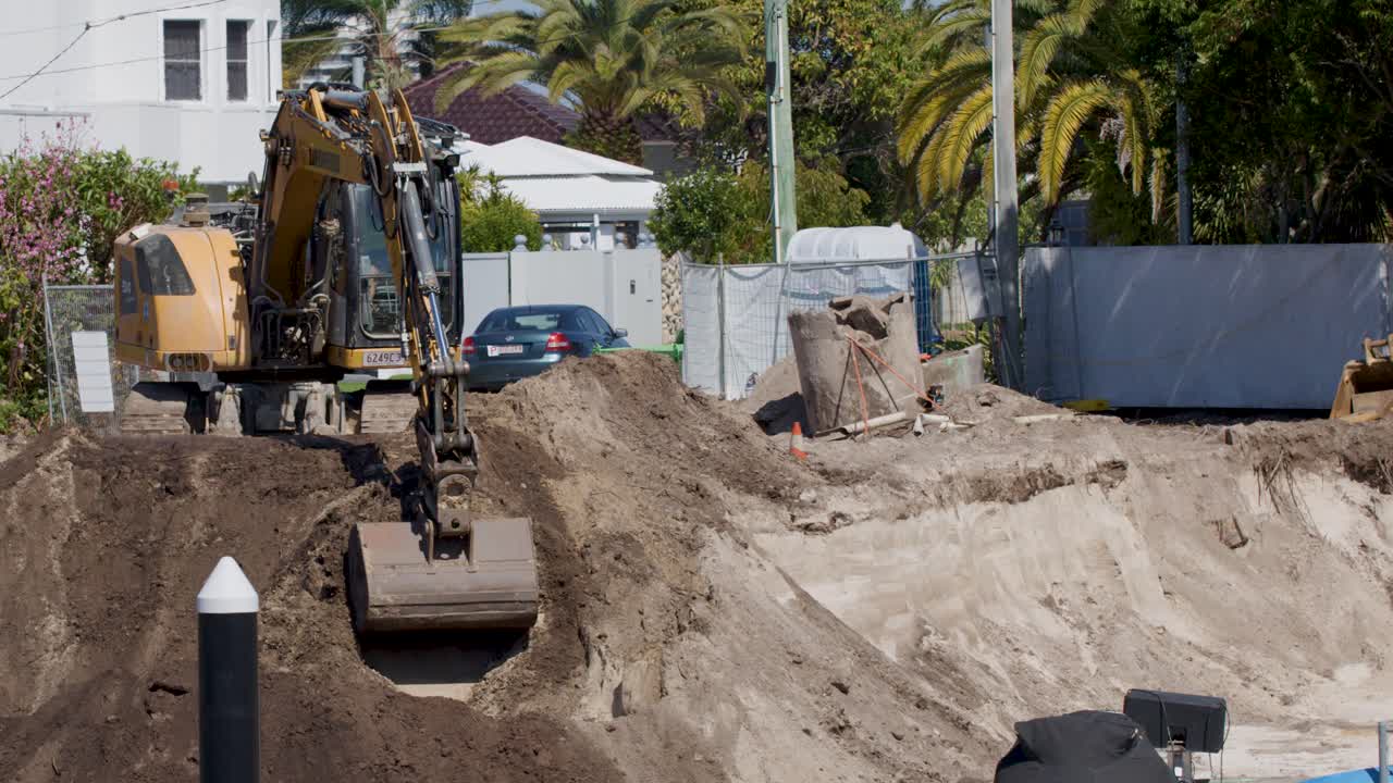 Excavator operates in bright daylight, digging earth for house redevelopment in suburban neighborhood