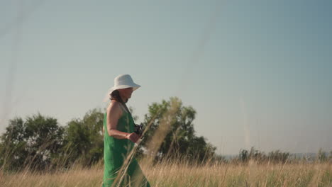 side view of confident woman in green dress and white sunhat walking through tall grass with camera in hand, surrounded by golden field under clear sky