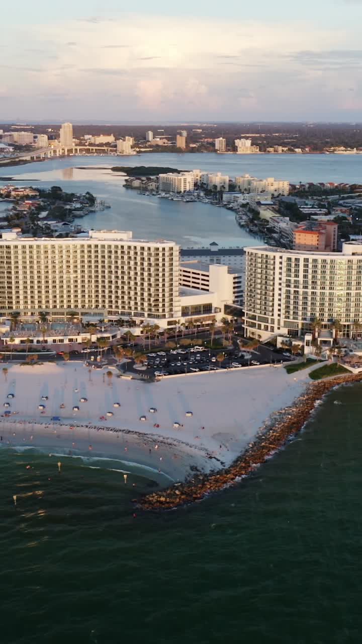 A wide sandy beach meets emerald waters as tall hotels overlook the coast, with a backdrop of a sprawling city skyline, bridges, and a calm bay