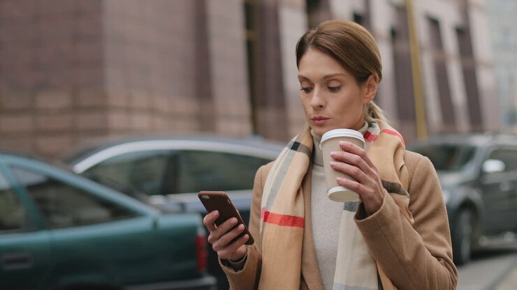 Caucasian charming businesswoman wearing coat and scarf walking on the street while drinking hot coffee and texting on the smartphone