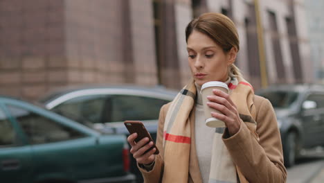 encantadora mujer de negocios caucásica con abrigo y bufanda caminando por la calle mientras bebe café caliente y envía mensajes de texto en el teléfono inteligente