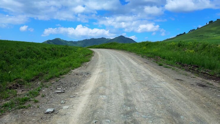 Una vista panoramica di una strada sterrata tortuosa, circondata da rigogliose colline verdi e un cielo azzurro e limpido con soffici nuvole bianche.