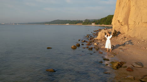 Caucasian-Woman-in-Dress-Enjoys-Stunning-Sunrise-At-Orlowo-Rocky-Beach-on-Calm-Summer-Morning-Standing-Seafront---Aerial-Flyover