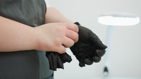 Close-up view of beautician adjusting black glove while wearing dark apron, standing in white lit salon with blurred background and lamp, preparing for professional client session