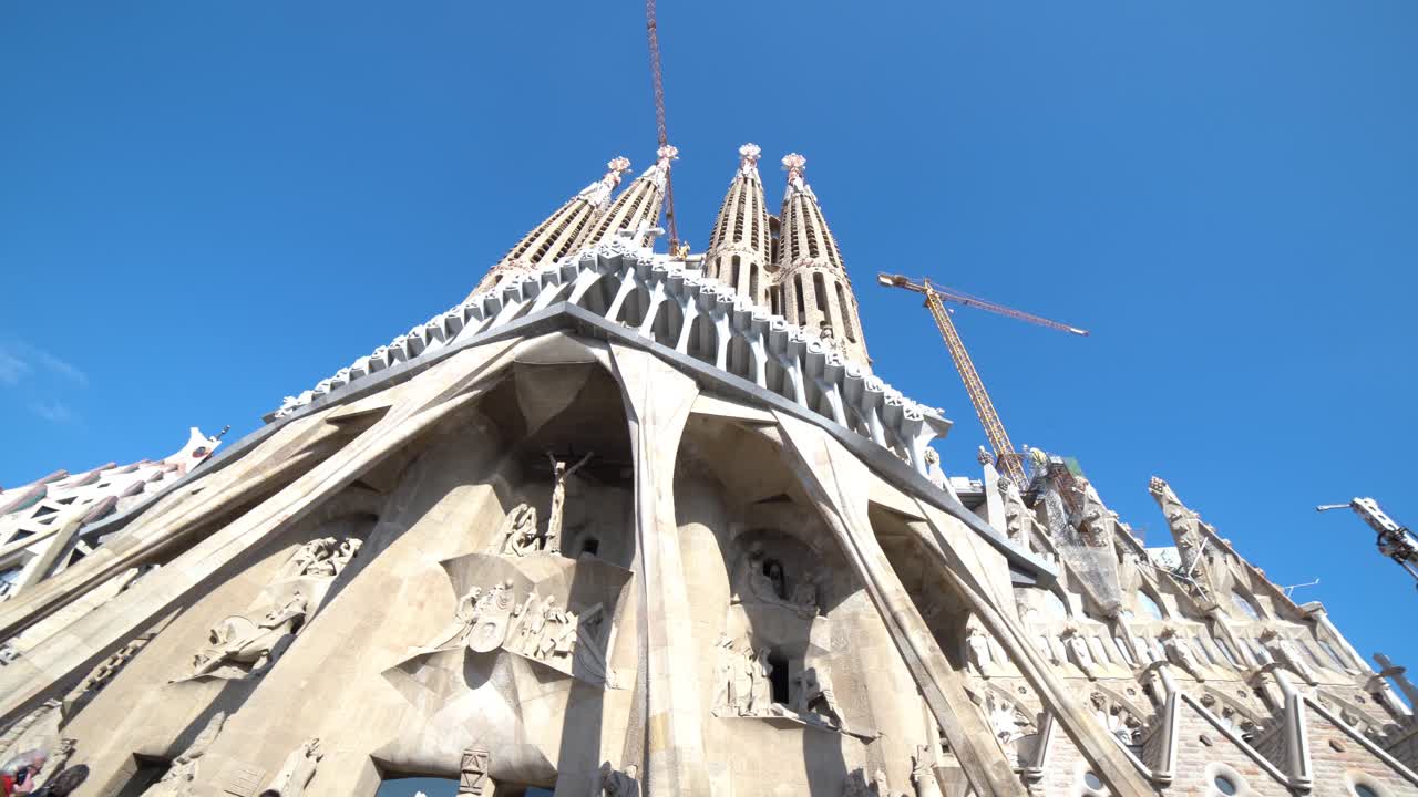Smooth wide upward look at the Christ Facade with blue sky at La Sagrada Familia with construction cranes, Barcelona. Wide pan from left to the right