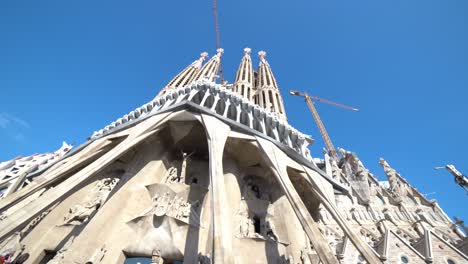 smooth wide upward look at the christ facade with blue sky at la sagrada familia with construction cranes, barcelona. wide pan from left to the right